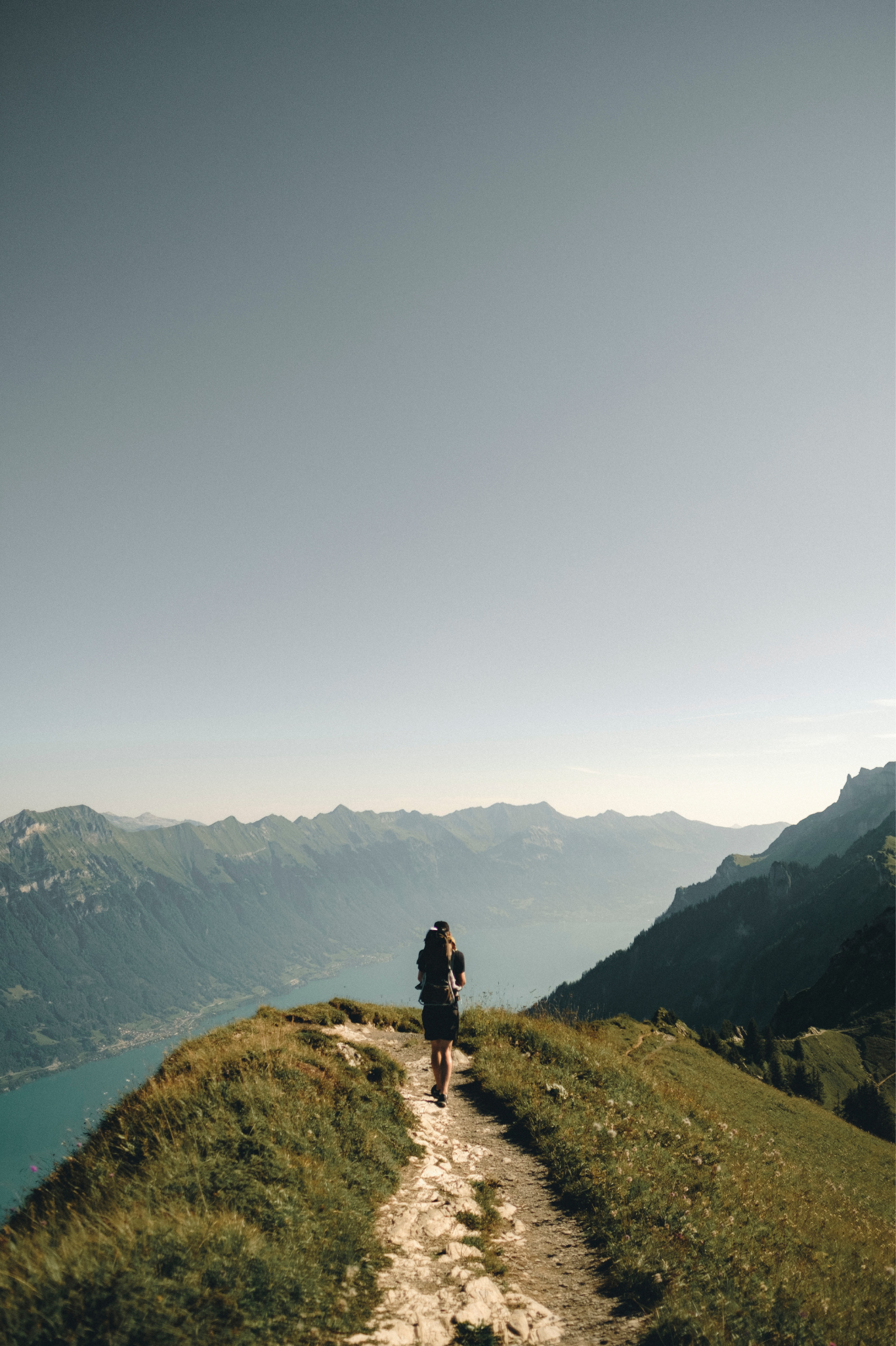 Zu sehen ist eine Person, die bei sonnigem Wetter auf einem Bergrücken wandert. Im Hintergrund zu sehen ist ein weiter Ausblick mit einer Gebirgskette und einem Fluss